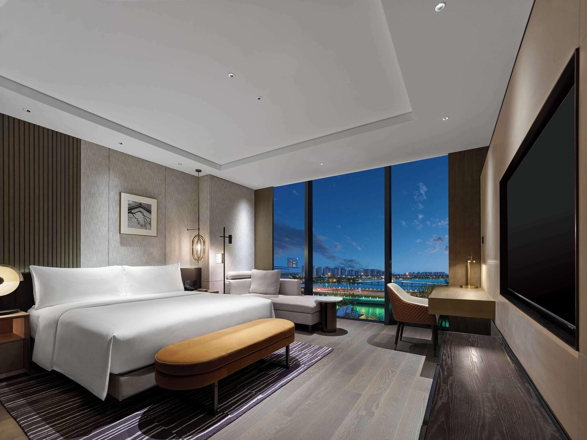 A modern hotel room with a large bed, a seating area, and a city view through large windows.