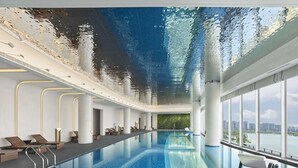 An indoor swimming pool with clear blue water, a ceiling that mimics water reflections, and a view of a cityscape through large windows. - Hilton Changshu (Suzhou)