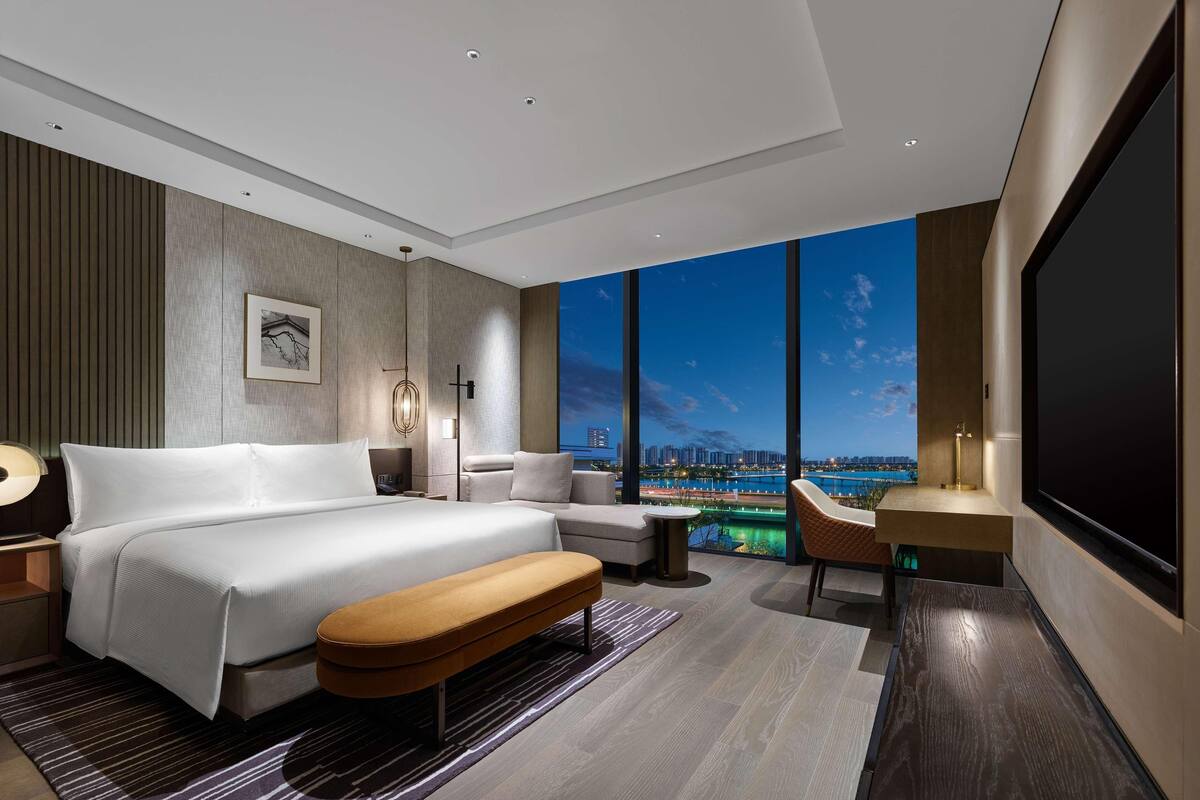 A modern hotel room with a large bed, a seating area, a desk, and a large window offering a city view.