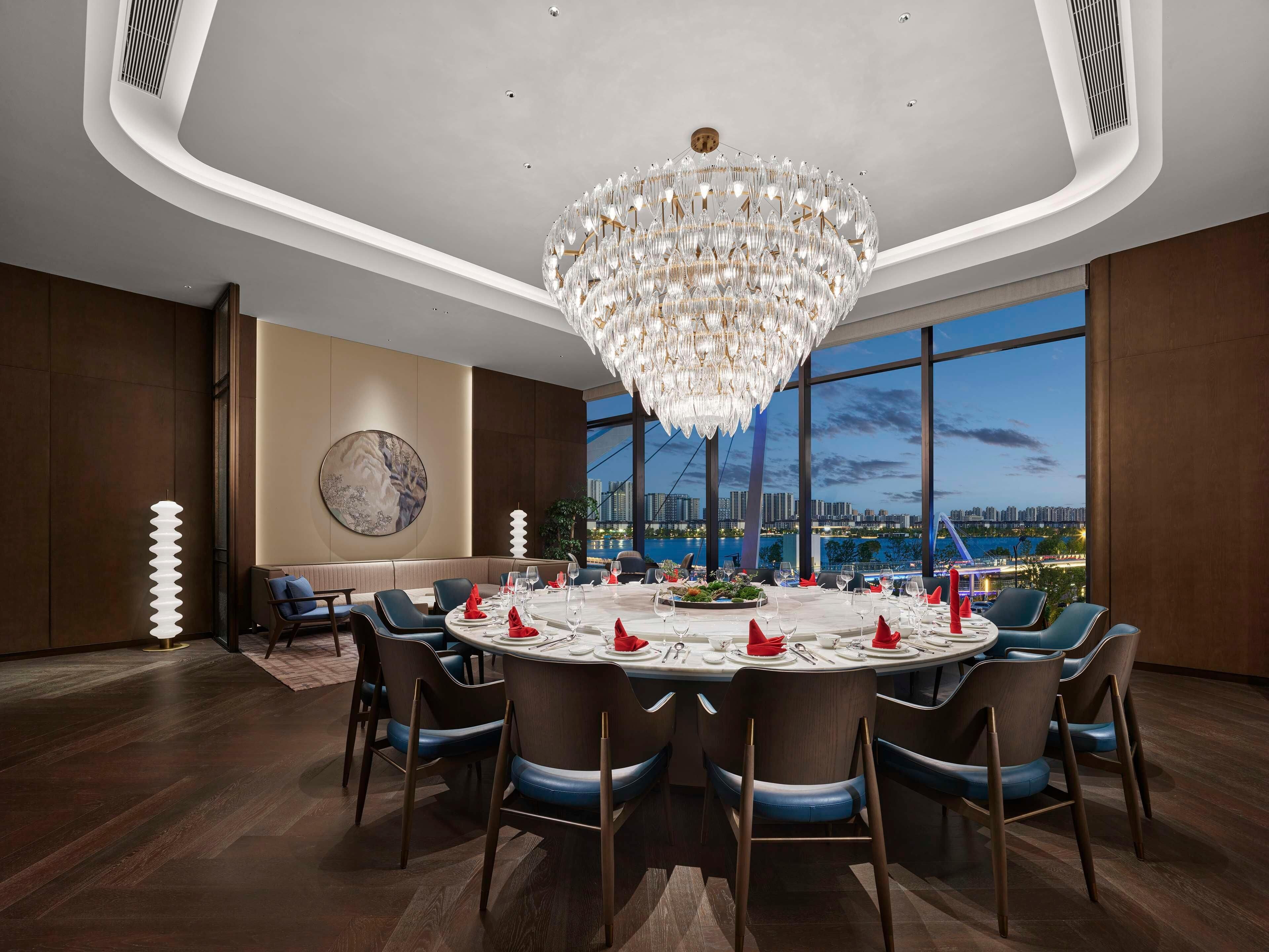 A modern dining room with a round table set for six, large windows offering a city view, and a prominent chandelier.