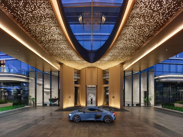 En modern hotellentrén med en blå sportbil parkerad framför, en glasfasad och ett tak med ett ljusmönster.