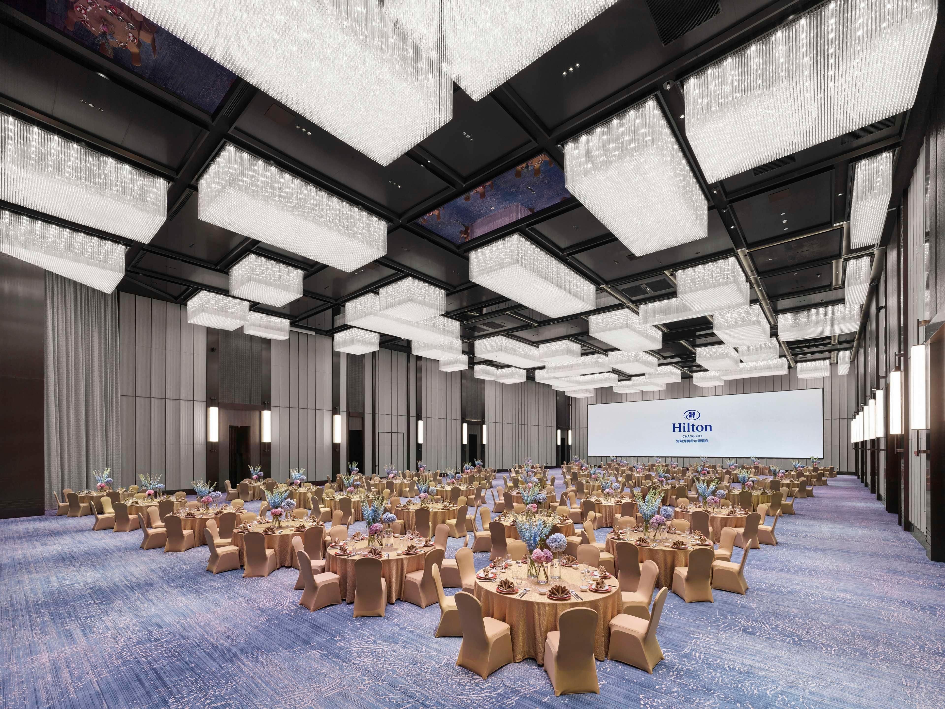 A large, well-lit conference room with round tables and chairs, a large screen displaying the Hilton logo, and a modern chandelier design.
