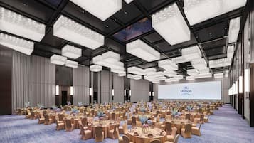 A large, well-lit conference room with round tables and chairs, a large screen displaying the Hilton logo, and a modern chandelier design.