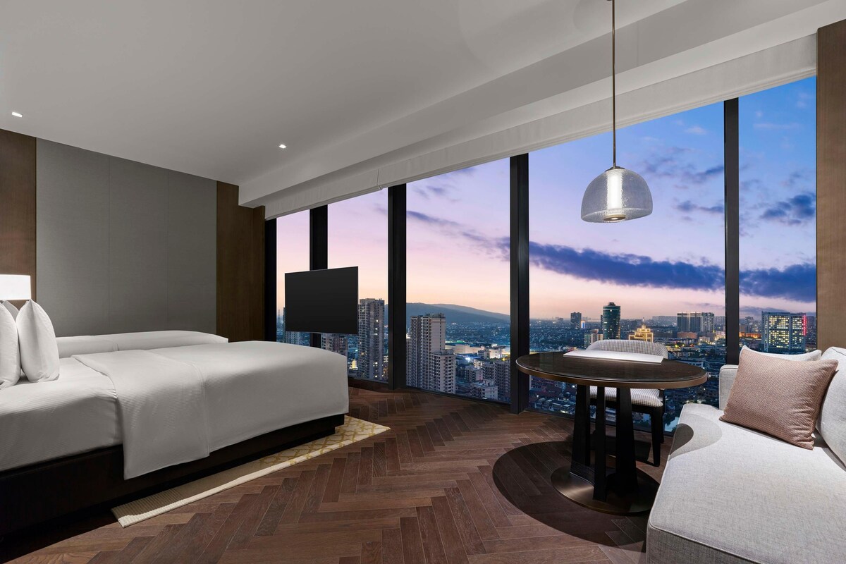 A modern hotel room with a large bed, a dining table, and a city view.
