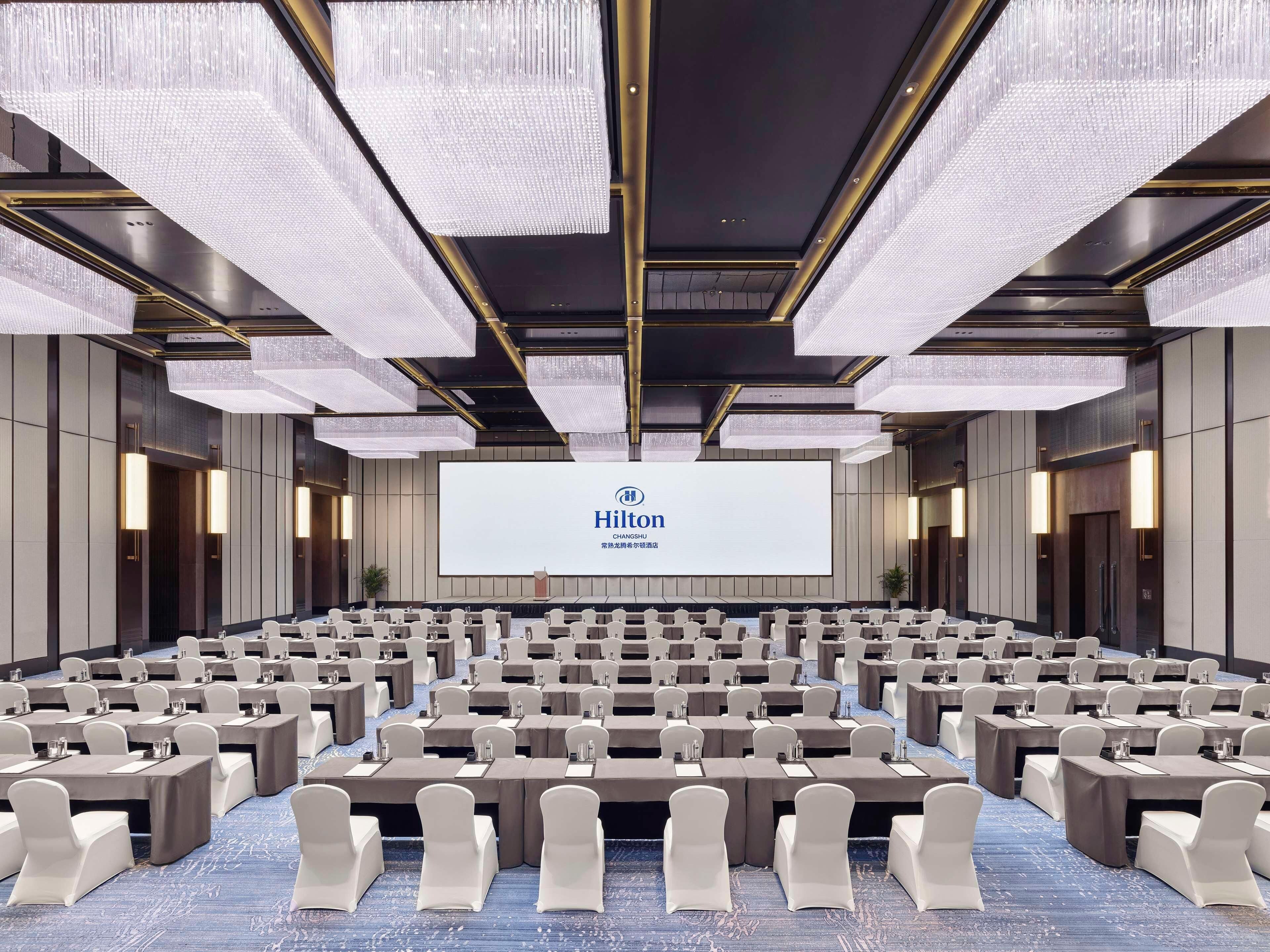 A conference room with rows of tables and chairs, a large screen displaying the Hilton logo, and a modern chandelier design.