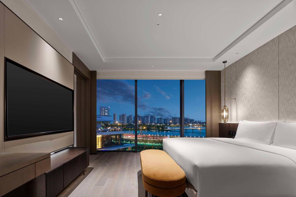 A modern hotel room with a large bed, a flat-screen TV, and a city view.