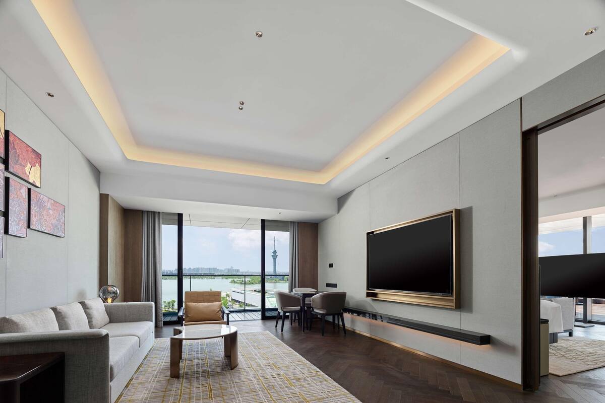 A modern living room with a large flat-screen TV, a sofa, a coffee table, and a view of a cityscape through large windows.