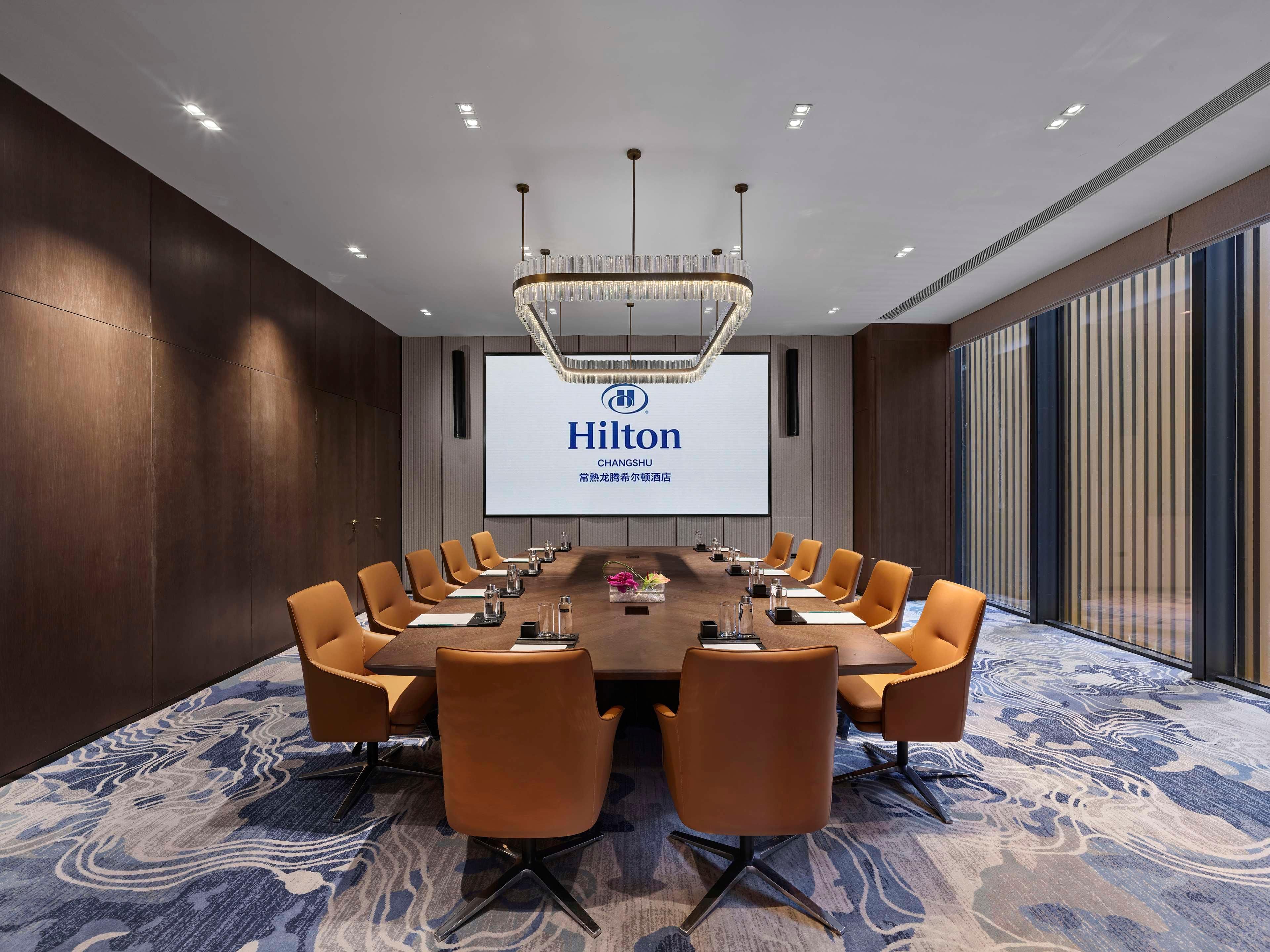 A conference room with a long table, multiple chairs, a large screen displaying 'Hilton CHANGSHU', and a modern chandelier.