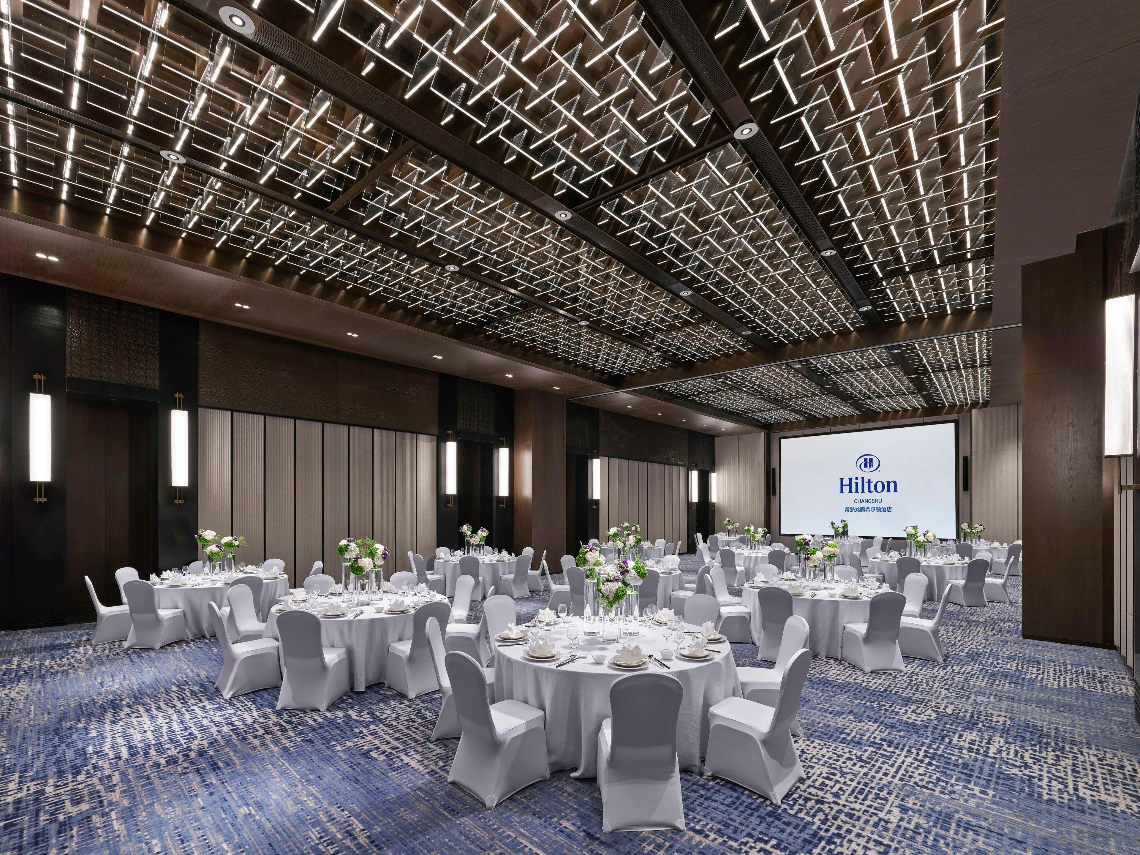 A conference room with multiple round tables, white chairs, and a large screen displaying the Hilton logo.