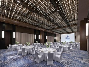 A conference room with multiple round tables, white chairs, and a large screen displaying the Hilton logo. - Hilton Changshu (Suzhou)