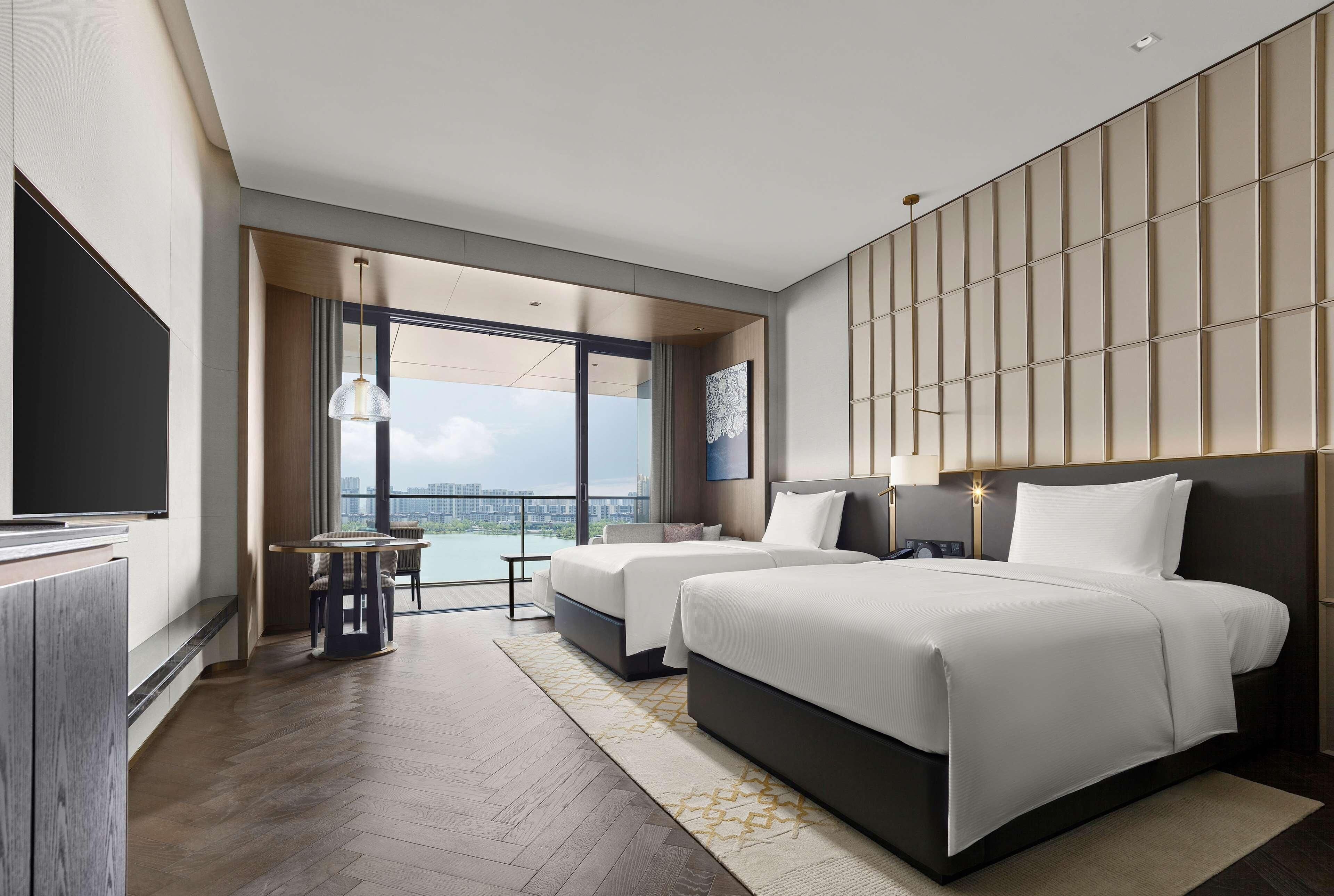 A modern hotel room with two beds, a TV, a balcony with a city view, and a large window.