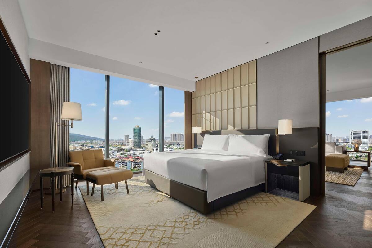 A modern hotel room with a large bed, a seating area with a chair and a small table, a nightstand with a lamp, and a large window offering a city view.