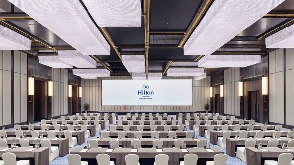 A large conference room with rows of tables and chairs, a large screen displaying the Hilton logo, and a modern ceiling design. - Hilton Changshu (Suzhou)