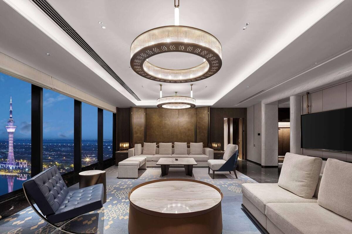 A modern hotel room with a large window offering a city view, a round coffee table, a sofa, and a TV.