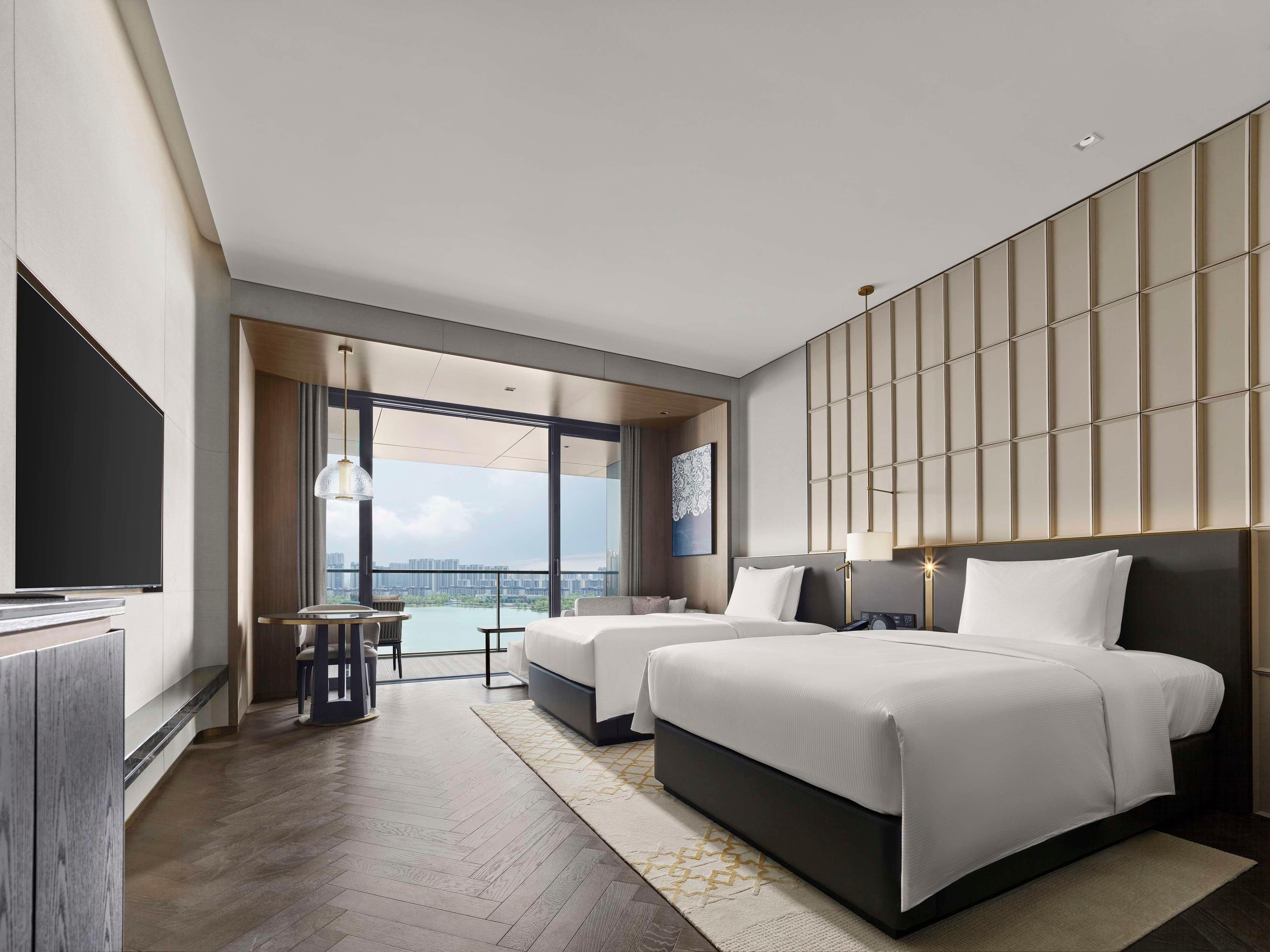 A modern hotel room with two beds, a large window with a city view, a flat-screen TV, and a small table with chairs.
