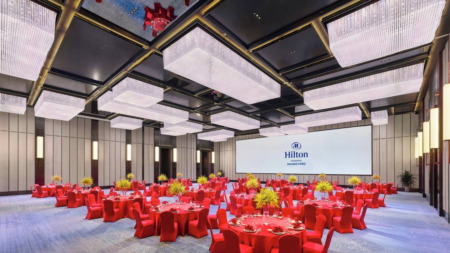 A spacious conference room with round tables and red chairs, a large screen displaying the Hilton logo, and a modern ceiling design.
