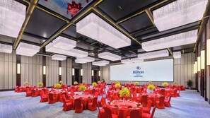 Een ruime congreszaal met ronde tafels en rode stoelen, een groot scherm waarop het Hilton logo staat afgebeeld, en een modern plafondontwerp.