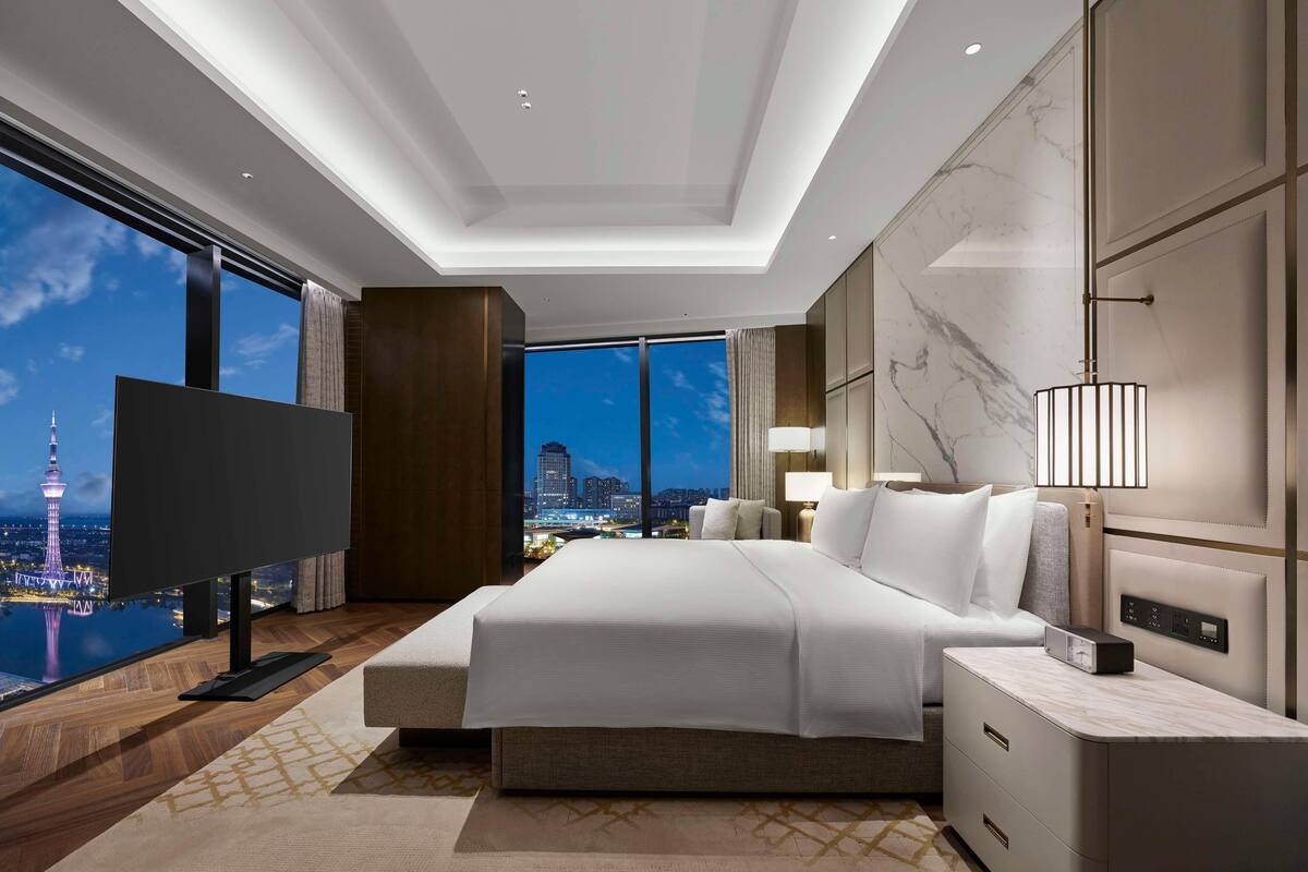 A modern hotel room with a large bed, a TV, and a view of a cityscape at night.