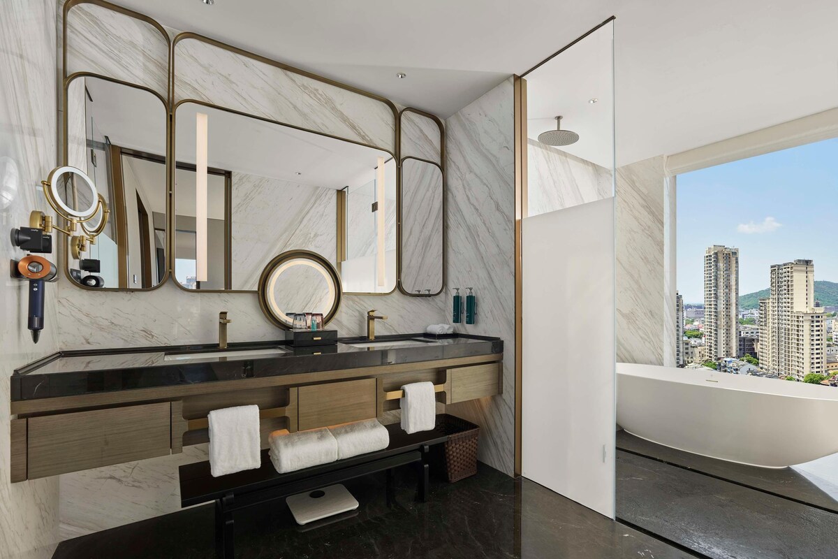 A modern bathroom with a marble vanity, multiple mirrors, a round light, and a view of tall buildings.