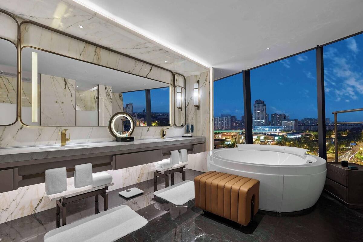 A modern bathroom with a large mirror, two sinks, a bathtub, and a city view.