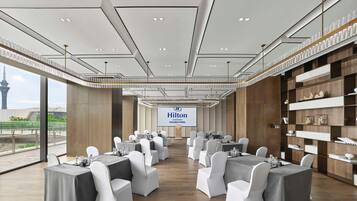 Une salle de conférence dotée de plusieurs tables et chaises, d’un grand écran affichant le nom « Hilton Changshu », et offrant une vue sur une tour à travers de grandes fenêtres.