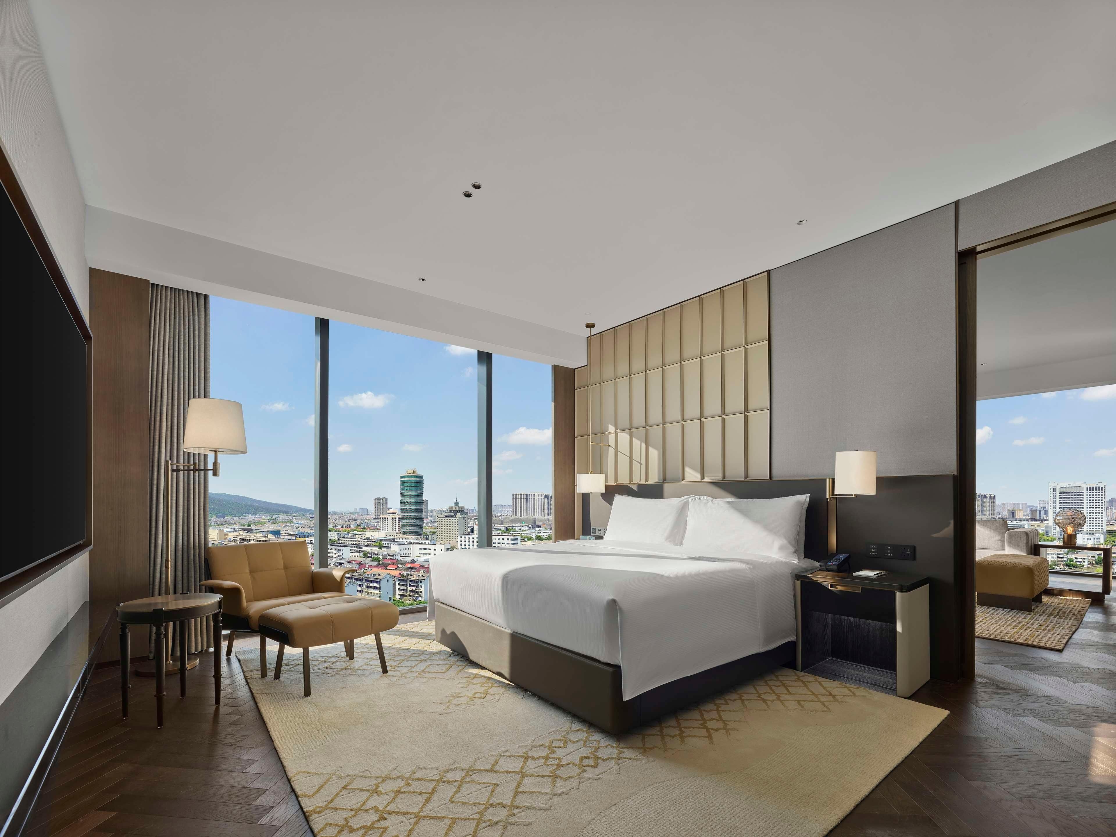 A modern hotel room with a large bed, a seating area with a chair and a small table, a nightstand with a lamp, and a large window offering a city view.