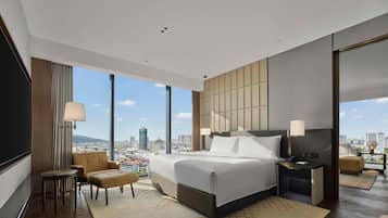 A modern hotel room with a large bed, a seating area with a chair and a small table, a nightstand with a lamp, and a large window offering a city view.