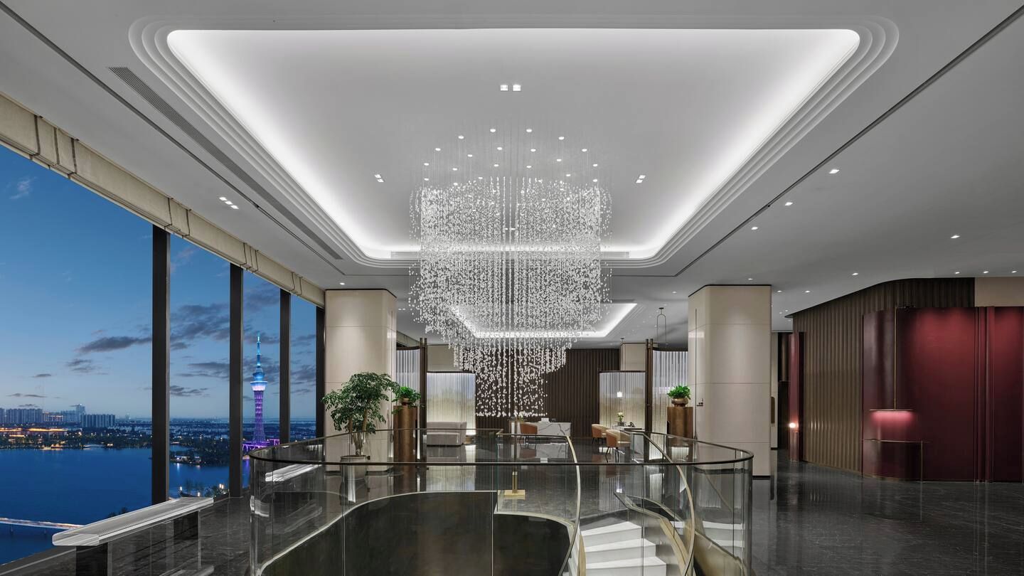 A modern hotel lobby with a large chandelier, a glass staircase, and a view of a city skyline.
