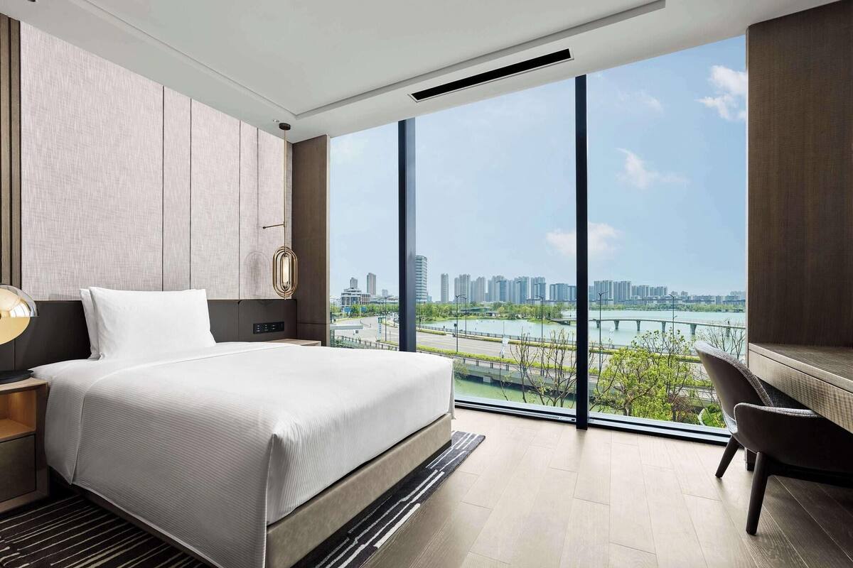 A modern hotel room with a large bed, a desk, and a view of a cityscape and river.