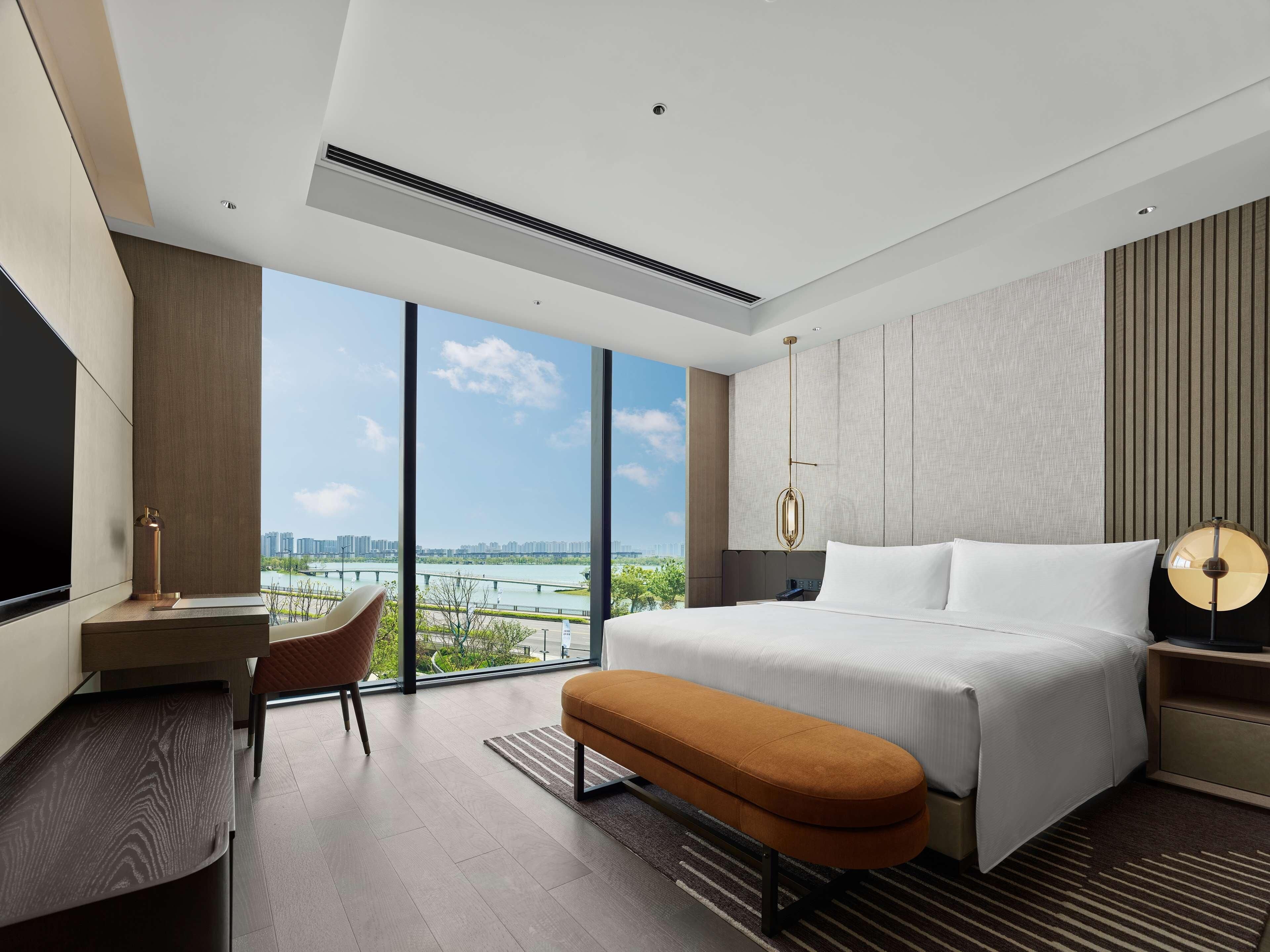 A modern hotel room with a large bed, a desk, a chair, and a view of a cityscape through large windows.