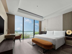 A modern hotel room with a large bed, a desk, a chair, and a view of a cityscape through large windows. - Hilton Changshu (Suzhou)