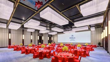 Une grande salle de conférence avec des tables rondes, des chaises rouges et un écran affichant le logo de la chaîne Hilton.