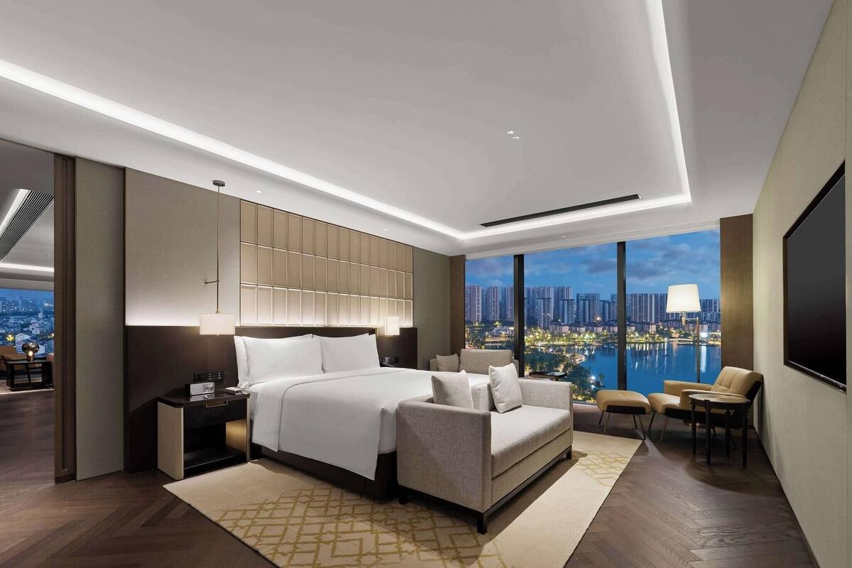 A modern hotel room with a large bed, a sofa, a TV, and a view of a cityscape and water.