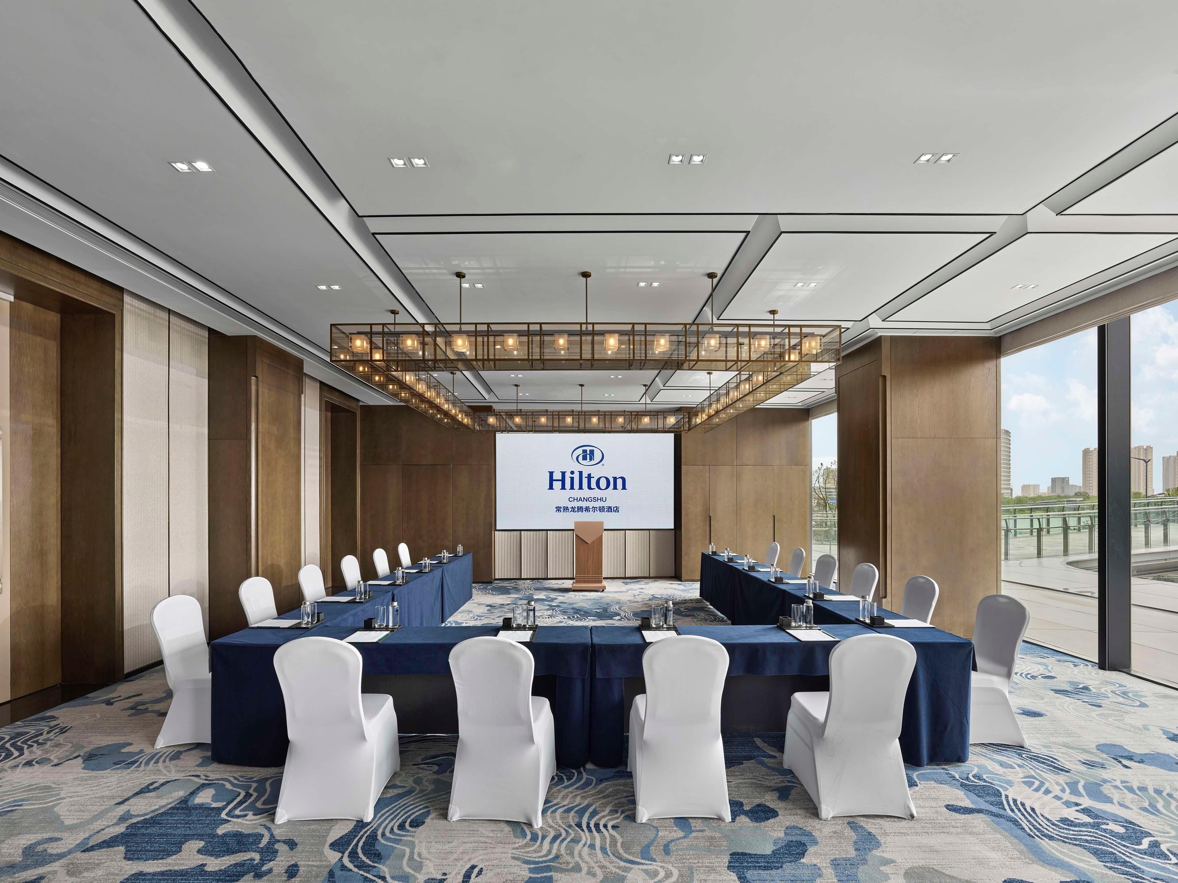 A conference room with a large screen displaying the Hilton logo, white chairs, and a blue carpet.
