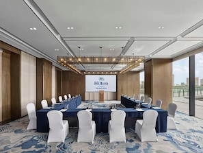 A conference room with a large screen displaying the Hilton logo, white chairs, and a blue carpet. - Hilton Changshu (Suzhou)