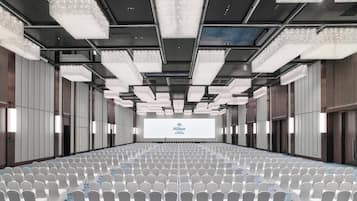 A large, empty conference room with rows of white chairs, a large screen at the front, and modern chandeliers.