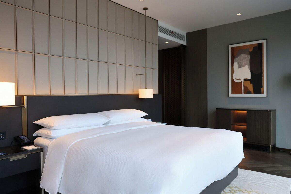 Een moderne hotelkamer met een groot bed, een nachtkastje met een lamp, een wandairconditioner en een lijst met een kunstwerk boven het bed.