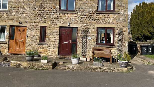 Une maison en pierre avec une porte en bois, une porte rouge et un banc à l’extérieur.