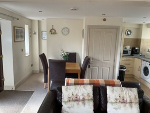 A living room with a black leather sofa, a wooden dining table with chairs, a kitchen area with appliances, and a door with a white frame. - Beautiful 2-bed Cottage in Grewelthorpe , Masham (Ripon)
