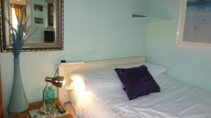 Een netjes opgemaakt bed met witte lakens en een enkel paars kussen, een bedlampje, een spiegel en een ingelijst foto aan de muur.