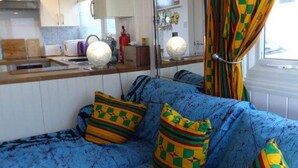 Een ruimtelijk ingerichte leefruimte met een kitchenette, een bank met opvallende kussens en een raam met gordijnen.