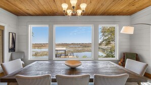Un comedor con mesa de madera, sillas a rayas, una araña y ventanas amplias con vistas a un paisaje natural.