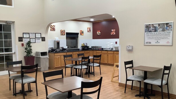 A dining area with wooden tables and chairs, a kitchen in the background, and a sign on the wall. - HomeTowne Studios by Red Roof Amarillo West (Amarillo)