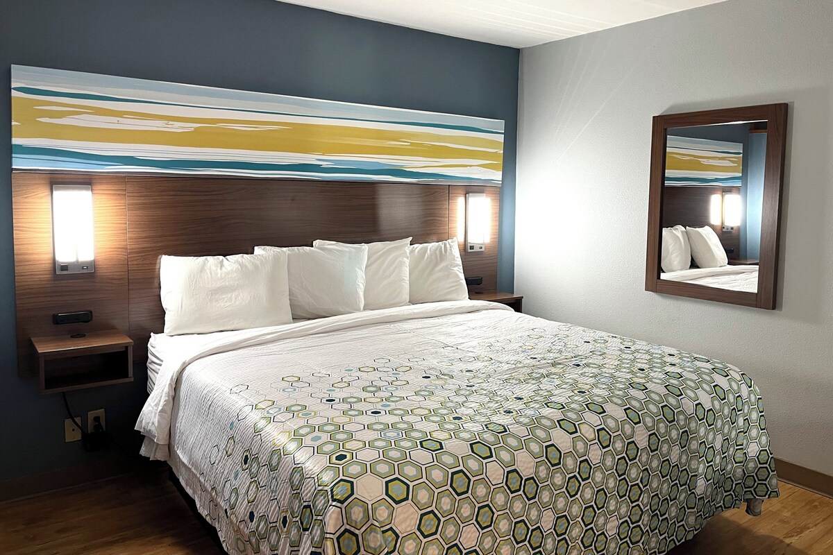 A hotel room with a large bed, a nightstand, a wall-mounted mirror, and a painting above the bed.