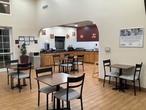 A hotel dining area with wooden tables and chairs, a kitchen in the background, and a framed informational poster on the wall. - HomeTowne Studios by Red Roof Amarillo West (Amarillo)
