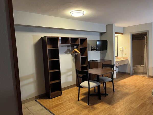 Habitación pequeña con suelo de madera, una mesita con dos sillas, un televisor montado en la pared y un armario con estantes.