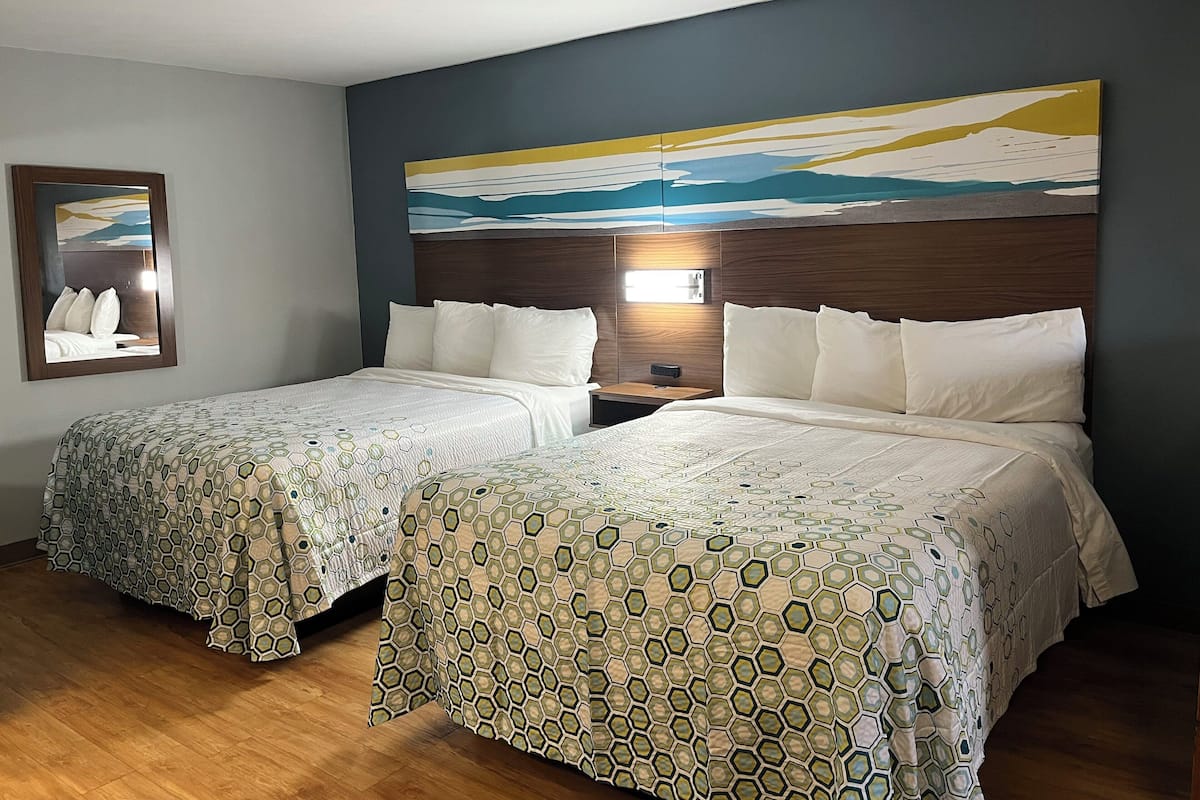 A hotel room with two beds, a nightstand, a wall-mounted lamp, and a framed picture on the wall.