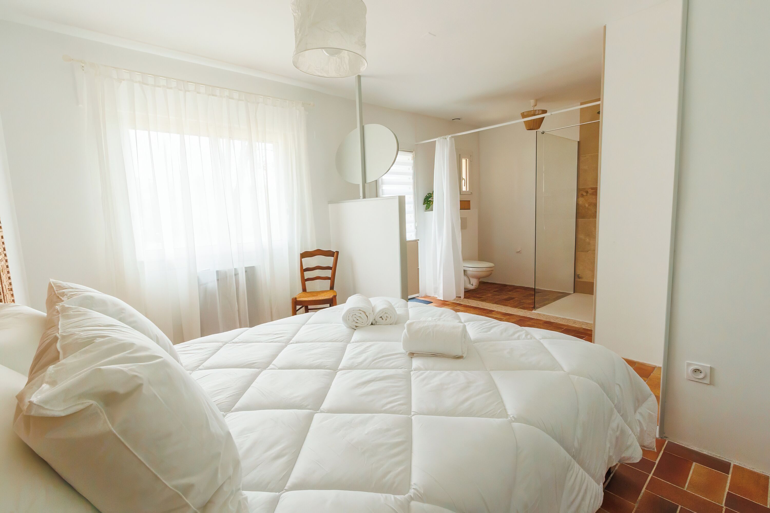 A bedroom with a large bed, a chair, a mirror, and a bathroom visible through an open door.