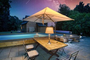 Ein Holztisch am Pool mit einer Lampe, umgeben von Stühlen, unter einem großen Sonnenschirm.
