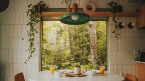 Ruokailutila, jossa on valkoinen pöytä, puiset tuolit ja suuri ikkuna, josta avautuu näkymä metsään.